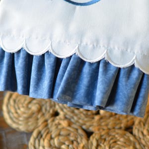 May include: A white fabric towel featuring a large embroidered navy blue letter S. The bottom edge of the towel has a scalloped hemline with a gathered, blue patterned fabric ruffle attached underneath the scalloped border.
