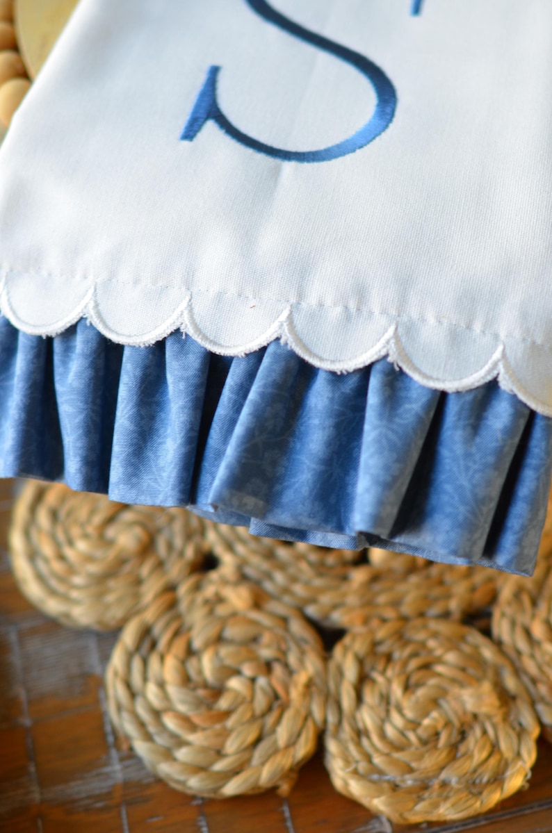 May include: A white fabric towel featuring a large embroidered navy blue letter S. The bottom edge of the towel has a scalloped hemline with a gathered, blue patterned fabric ruffle attached underneath the scalloped border.