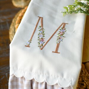 May include: A cream-colored hand towel featuring an embroidered letter M in a brown outline, decorated with small blue, pink, and green floral accents. The bottom edge has a scalloped hem and a ruffled trim in a light brown and white gingham pattern.