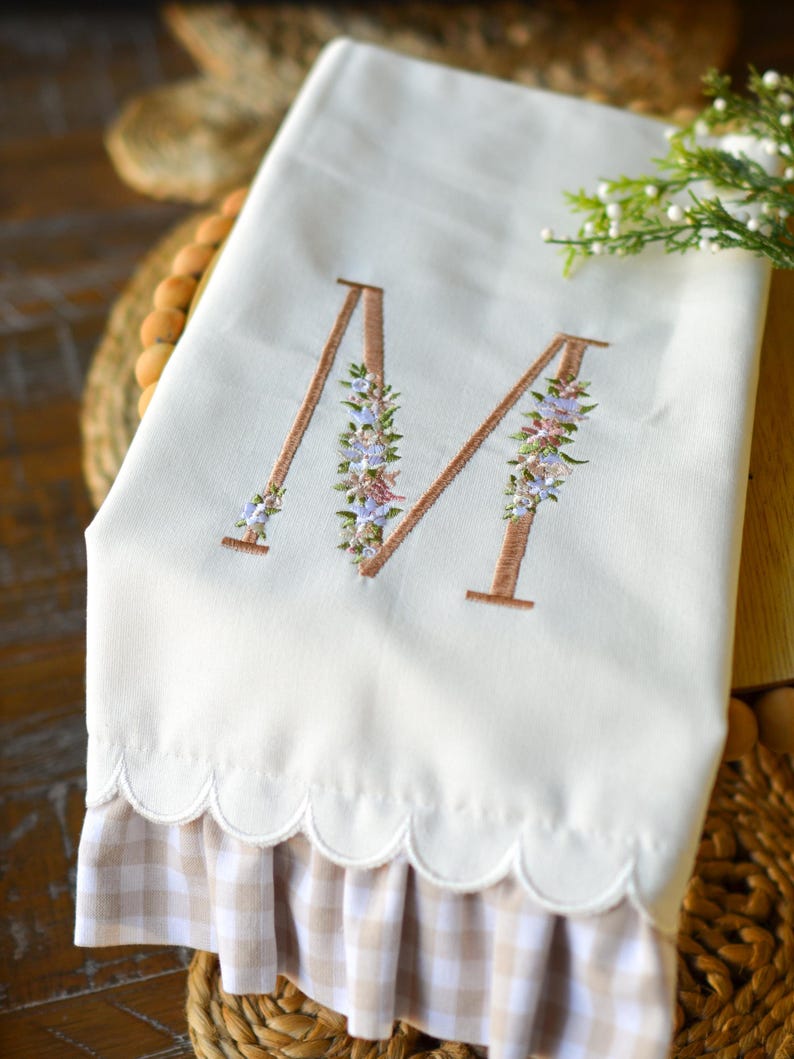 May include: A cream-colored hand towel featuring an embroidered letter M in a brown outline, decorated with small blue, pink, and green floral accents. The bottom edge has a scalloped hem and a ruffled trim in a light brown and white gingham pattern.