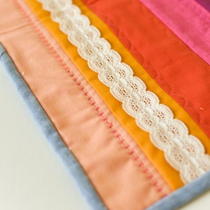 May include: A colorful, handmade quilt with a patchwork design. The quilt features strips of orange, pink, and yellow fabric, accented with white lace trim. The edges are finished with a blue border and pink stitching.