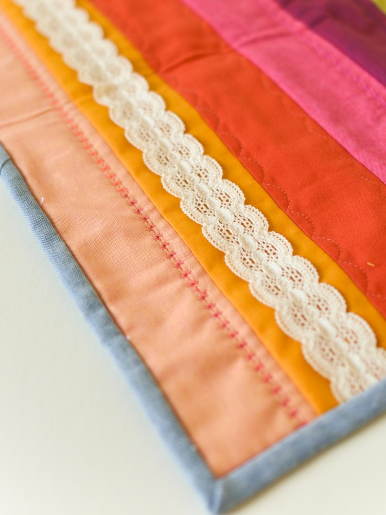 May include: A colorful, handmade quilt with a patchwork design. The quilt features strips of orange, pink, and yellow fabric, accented with white lace trim. The edges are finished with a blue border and pink stitching.