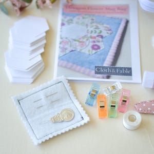 May include: A craft kit with a "Dragon Flower Mug Rug" pattern, white paper shapes, colorful clips, and a needle holder. The kit includes a spool of white tape and a pink and white polka dot clip.