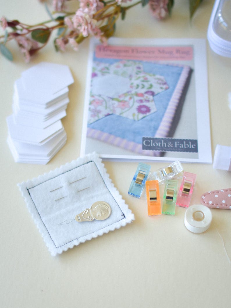 May include: A craft kit with a "Dragon Flower Mug Rug" pattern, white paper shapes, colorful clips, and a needle holder. The kit includes a spool of white tape and a pink and white polka dot clip.
