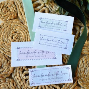 Op de afbeelding: Vier witte kaarten met de tekst "handmade with love" in een decoratief lettertype, elk met een andere naam en jaar. De kaarten liggen op een geweven oppervlak met groen lint en bladeren.