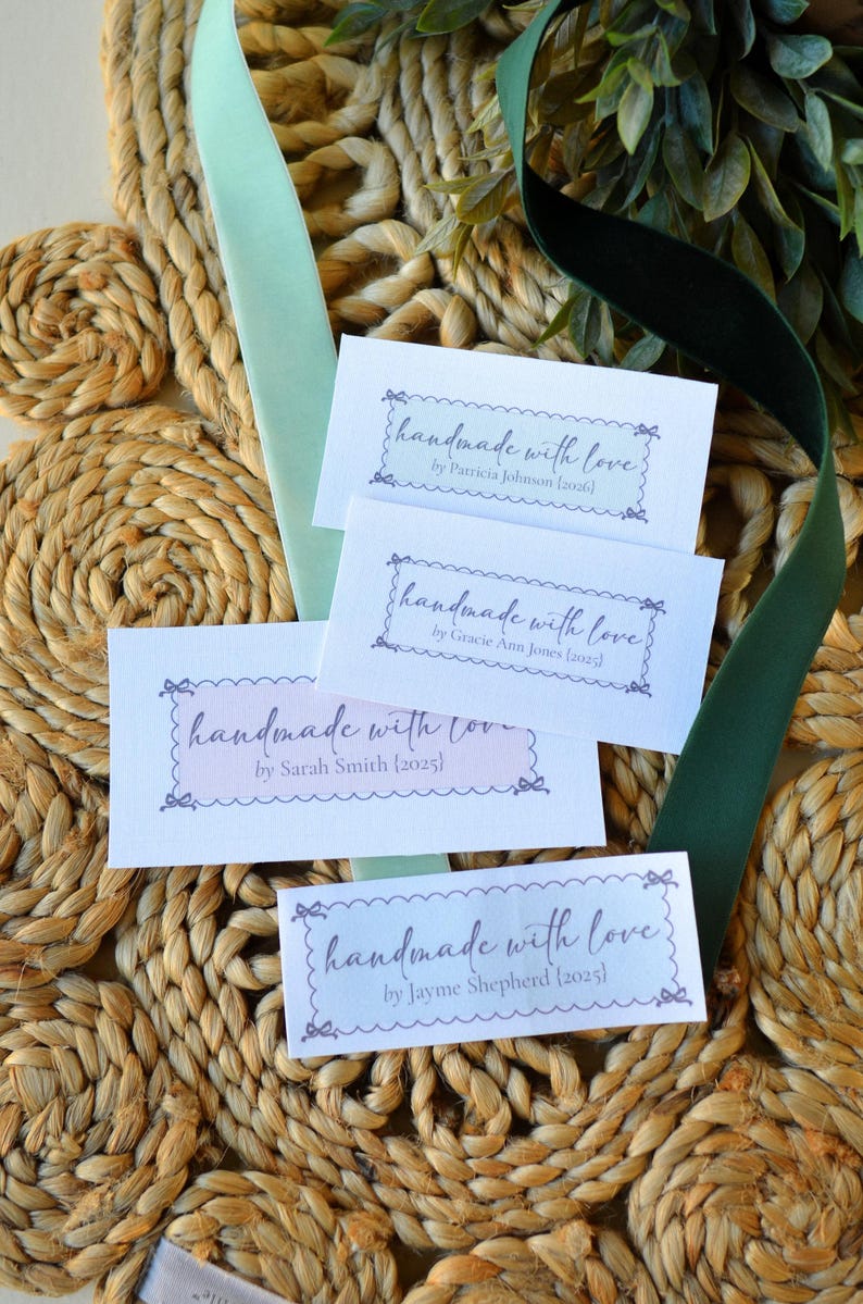Op de afbeelding: Vier witte kaarten met de tekst "handmade with love" in een decoratief lettertype, elk met een andere naam en jaar. De kaarten liggen op een geweven oppervlak met groen lint en bladeren.