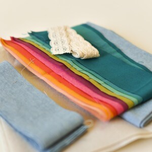 May include: A collection of colorful fabric scraps in shades of blue, green, yellow, orange, and pink. A piece of cream-colored lace is placed on top of the fabric. The fabrics are arranged on a white surface.