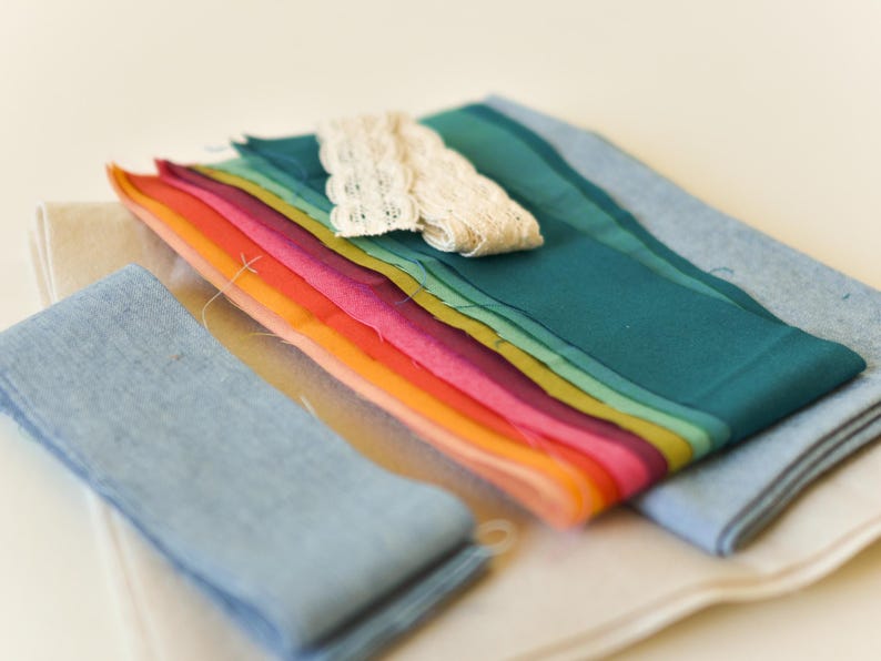 May include: A collection of colorful fabric scraps in shades of blue, green, yellow, orange, and pink. A piece of cream-colored lace is placed on top of the fabric. The fabrics are arranged on a white surface.