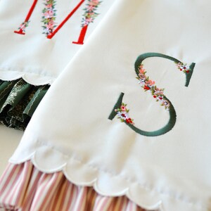 May include: Two white fabric towels featuring embroidered letters M and S. The letter M is red with floral accents, and the letter S is dark green with floral accents. Both towels have scalloped edges and bottom ruffles with striped patterns.