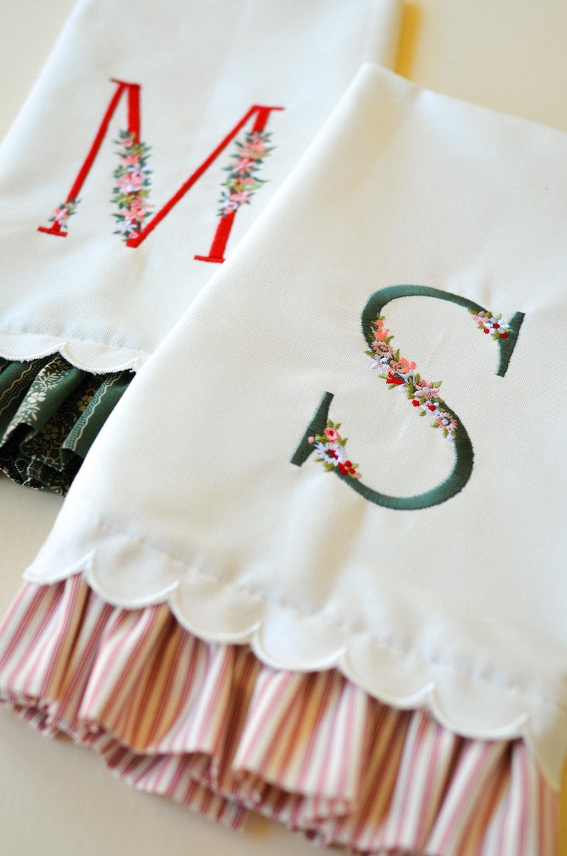 May include: Two white fabric towels featuring embroidered letters M and S. The letter M is red with floral accents, and the letter S is dark green with floral accents. Both towels have scalloped edges and bottom ruffles with striped patterns.