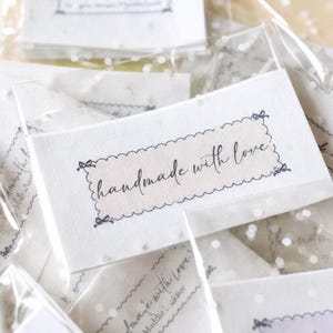 Op de afbeelding: Close-up van verschillende kleine, witte kaarten in transparante plastic verpakking. Elke kaart heeft de woorden "handmade with love" in cursiefschrift binnen een decoratieve rand. De kaarten zijn verspreid, wat een zachte, uitnodigende uitstraling cre&euml;ert.