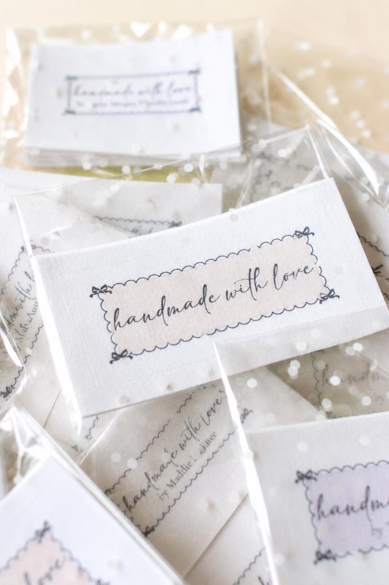 Op de afbeelding: Close-up van verschillende kleine, witte kaarten in transparante plastic verpakking. Elke kaart heeft de woorden "handmade with love" in cursiefschrift binnen een decoratieve rand. De kaarten zijn verspreid, wat een zachte, uitnodigende uitstraling cre&euml;ert.