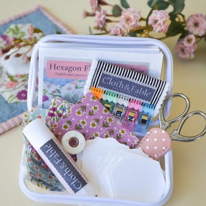 May include: A white zippered case filled with sewing supplies. Includes fabric scraps, paper pieces, scissors, thread, clips, and a tube of glue. The case contains items with the text "Cloth & Fable" and "Hexagon F".