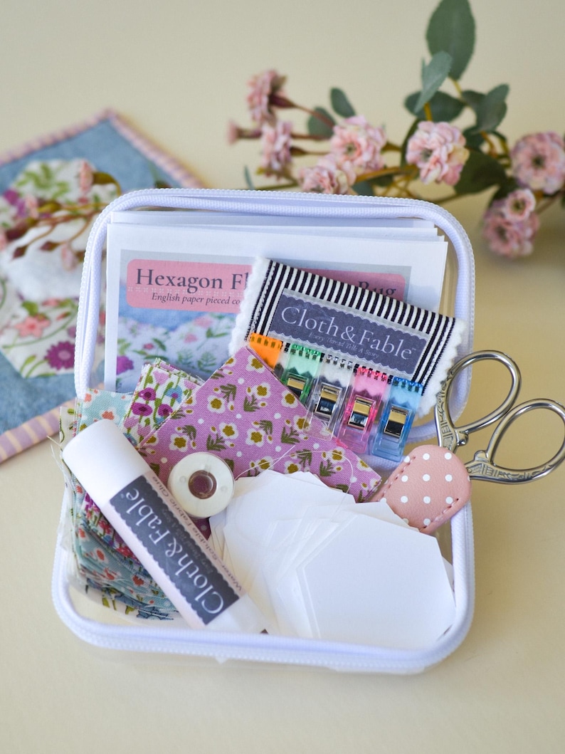 May include: A white zippered case filled with sewing supplies. Includes fabric scraps, paper pieces, scissors, thread, clips, and a tube of glue. The case contains items with the text "Cloth & Fable" and "Hexagon F".