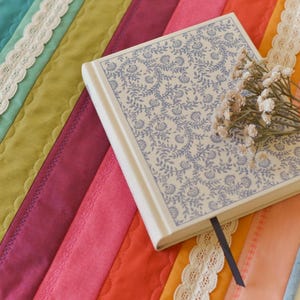 May include: A colorful, quilted placemat with a rainbow of hues, including teal, green, pink, and orange, is adorned with white lace trim. A floral-patterned book with a blue ribbon bookmark and a small bouquet of dried flowers rests on the placemat.