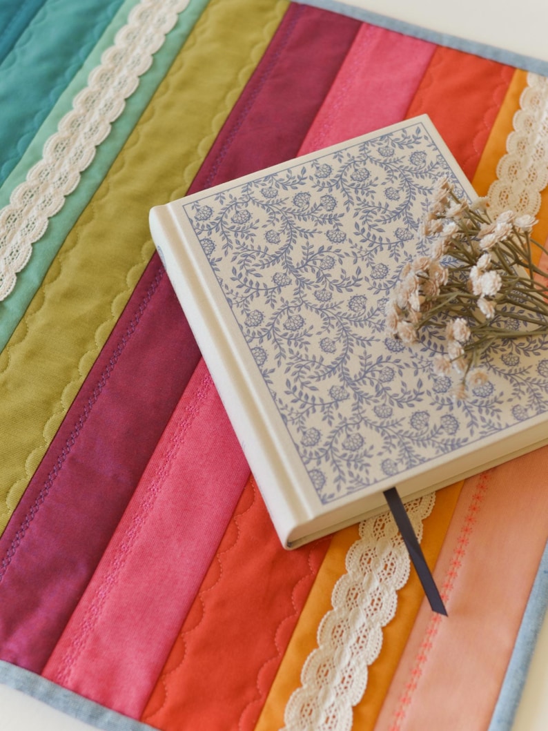 May include: A colorful, quilted placemat with a rainbow of hues, including teal, green, pink, and orange, is adorned with white lace trim. A floral-patterned book with a blue ribbon bookmark and a small bouquet of dried flowers rests on the placemat.