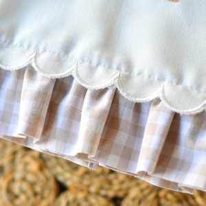 May include: A cream-colored fabric item featuring a scalloped bottom edge with a tan and white gingham ruffle trim. The upper portion displays a portion of an embroidered letter M decorated with small, colorful floral accents in pink and green.
