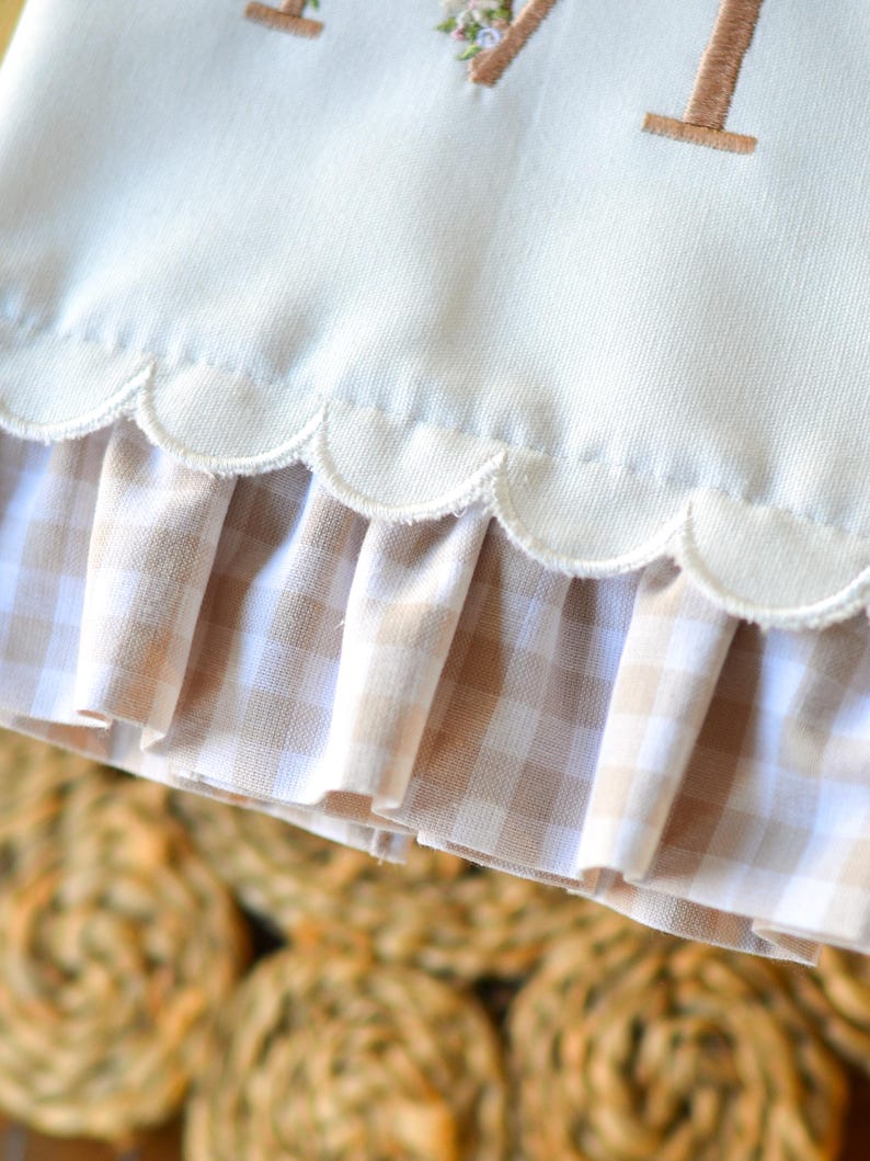 May include: A cream-colored fabric item featuring a scalloped bottom edge with a tan and white gingham ruffle trim. The upper portion displays a portion of an embroidered letter M decorated with small, colorful floral accents in pink and green.