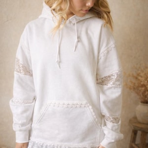 May include: White hoodie with lace accents on the sleeves, pocket, and hem. The hoodie has a drawstring and a front pocket. The sleeves and hem are trimmed with delicate lace. The model is wearing denim shorts.