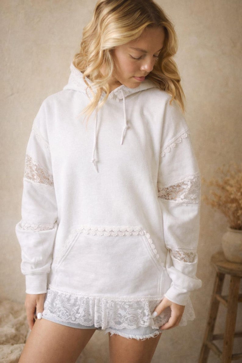 May include: White hoodie with lace accents on the sleeves, pocket, and hem. The hoodie has a drawstring and a front pocket. The sleeves and hem are trimmed with delicate lace. The model is wearing denim shorts.