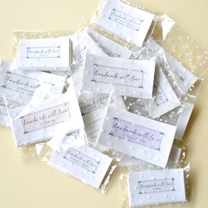 Op de afbeelding: Een verzameling kleine, witte kaarten met de tekst "handmade with love" in een sierlijk lettertype, verpakt in transparante, met stippen bedrukte plastic zakjes. De kaarten hebben een geschulpte rand.
