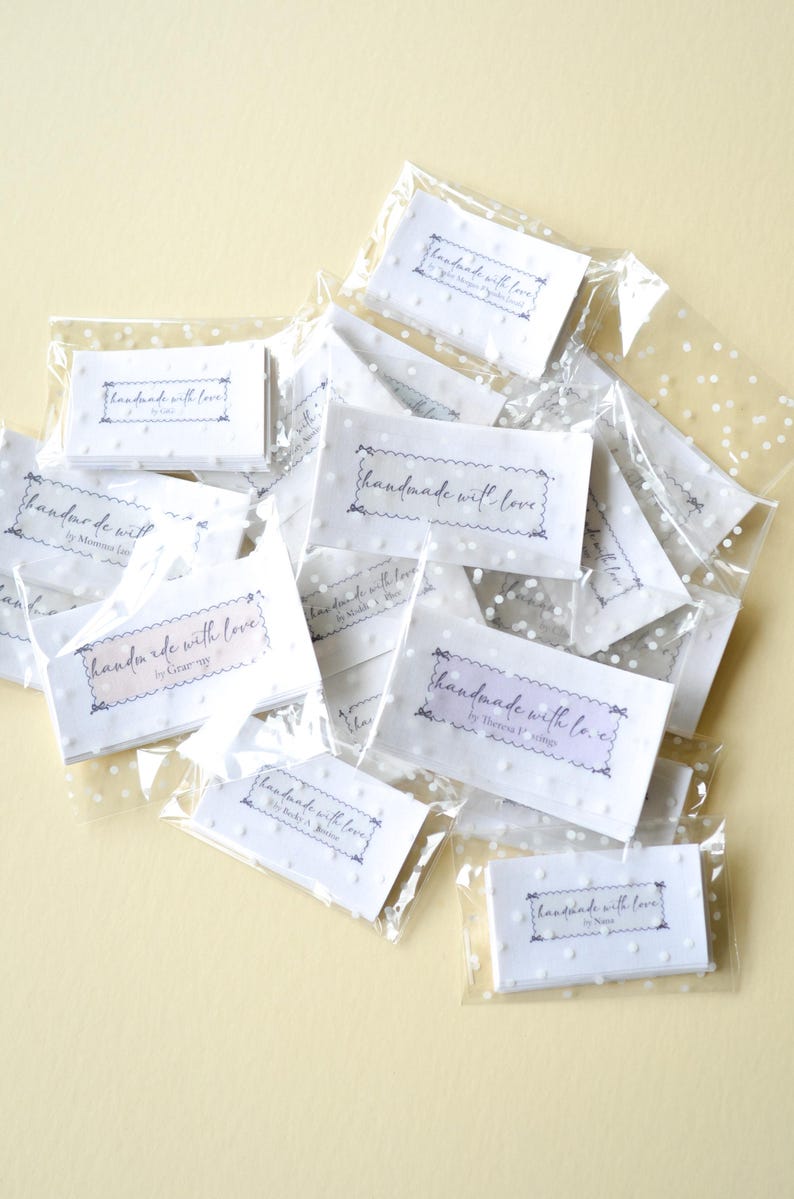 Op de afbeelding: Een verzameling kleine, witte kaarten met de tekst "handmade with love" in een sierlijk lettertype, verpakt in transparante, met stippen bedrukte plastic zakjes. De kaarten hebben een geschulpte rand.