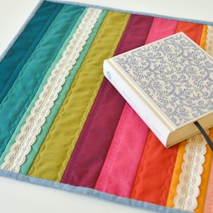 May include: A colorful, quilted placemat with vertical stripes in teal, green, pink, and orange, accented with white lace trim. A hardback book with a blue floral design rests on the mat, with a blue bookmark.