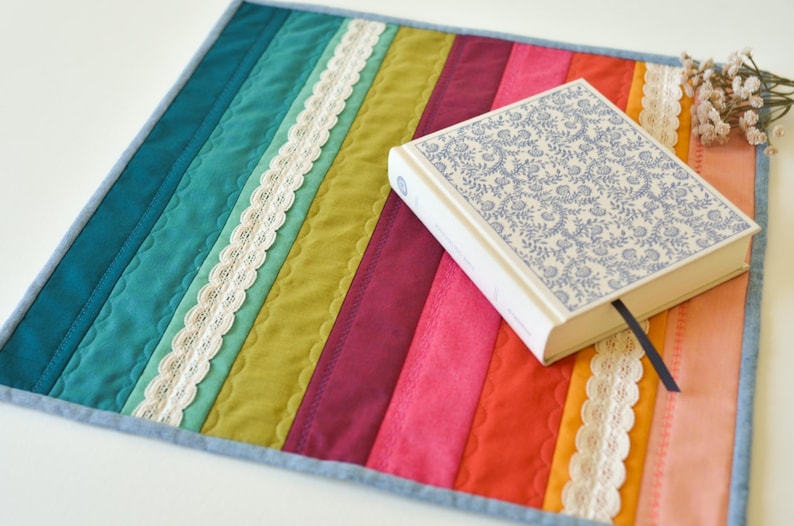 May include: A colorful, quilted placemat with vertical stripes in teal, green, pink, and orange, accented with white lace trim. A hardback book with a blue floral design rests on the mat, with a blue bookmark.