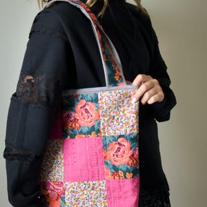Puede incluir: Un bolso tote patchwork con un estampado floral. La bolsa presenta una combinación de cuadrados de tela rosa y floral, con una correa a rayas. El estampado floral incluye grandes rosas rosas y flores más pequeñas.