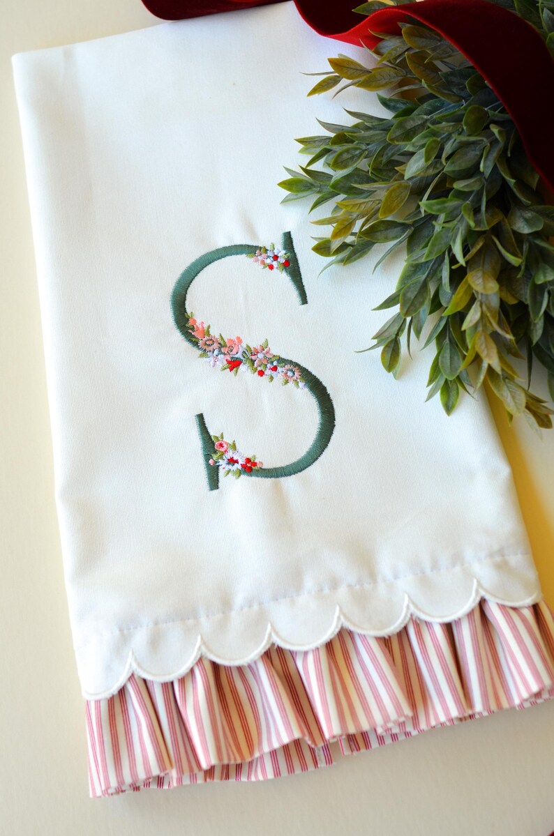 May include: A white hand towel featuring a dark green embroidered letter S decorated with small pink, red, and white floral accents. The bottom edge of the towel has a scalloped hem and a ruffled trim with a red and white vertical stripe pattern.