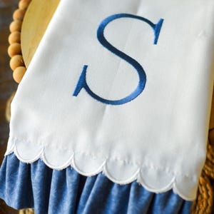 May include: A white hand towel featuring a large, embroidered navy blue letter S in the center. The bottom edge of the towel has a scalloped hemline and is finished with a gathered, ruffled blue fabric trim that has a subtle tonal pattern.