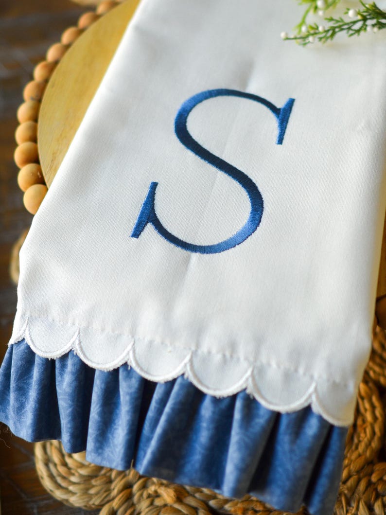 May include: A white hand towel featuring a large, embroidered navy blue letter S in the center. The bottom edge of the towel has a scalloped hemline and is finished with a gathered, ruffled blue fabric trim that has a subtle tonal pattern.