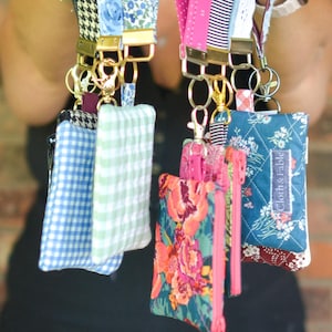 May include: A collection of colorful lanyards with matching small zippered pouches. The lanyards feature various patterns, including floral, gingham, and stripes. The pouches have coordinating fabrics and zippers, with gold-toned hardware.