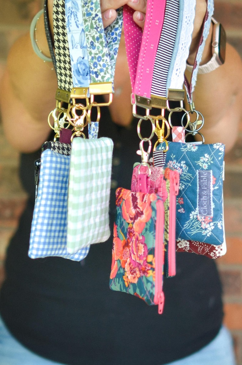 May include: A collection of colorful lanyards with matching small zippered pouches. The lanyards feature various patterns, including floral, gingham, and stripes. The pouches have coordinating fabrics and zippers, with gold-toned hardware.
