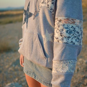 May include: A gray hoodie with star and floral patchwork details. The hoodie features a large dark blue star on the front and a white star on the pocket. The sleeves have floral and lace accents. The person is wearing a denim skirt.