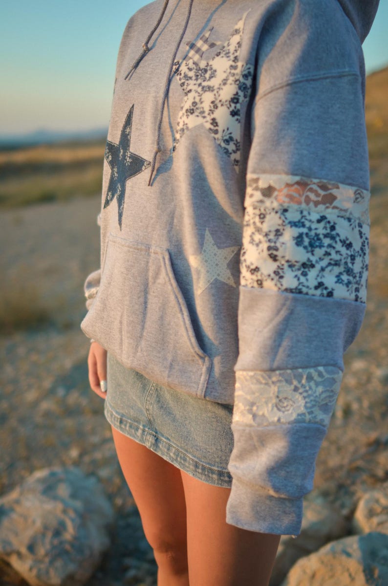 May include: A gray hoodie with star and floral patchwork details. The hoodie features a large dark blue star on the front and a white star on the pocket. The sleeves have floral and lace accents. The person is wearing a denim skirt.