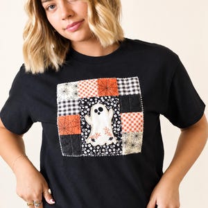 May include: A black short-sleeved t-shirt featuring a central square patch made of various fabric squares. The patch includes patterns of orange and black spiderwebs, black and white gingham, and floral prints, with a white ghost appliqué in the center.