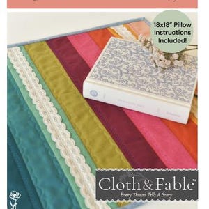 May include: A colorful Boho Stripe Mini Quilt with a striped design in teal, green, burgundy, pink, and orange. The quilt is accented with white lace trim. A book with a floral pattern and the text "Cloth & Fable" is on top of the quilt. Finished size is 18x18 inches.