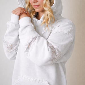 May include: A white hoodie with lace detailing on the hood, sleeves, and hem. The hoodie has a front pocket and a drawstring. The sleeves are long and feature lace inserts. The overall style is casual and feminine.