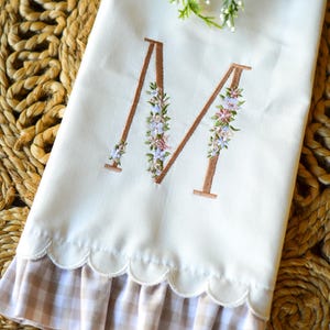 May include: A white hand towel featuring an embroidered letter M in a copper-colored thread, accented with small floral details in shades of pink, blue, and green. The bottom edge has a scalloped finish with a beige and white gingham ruffle trim.