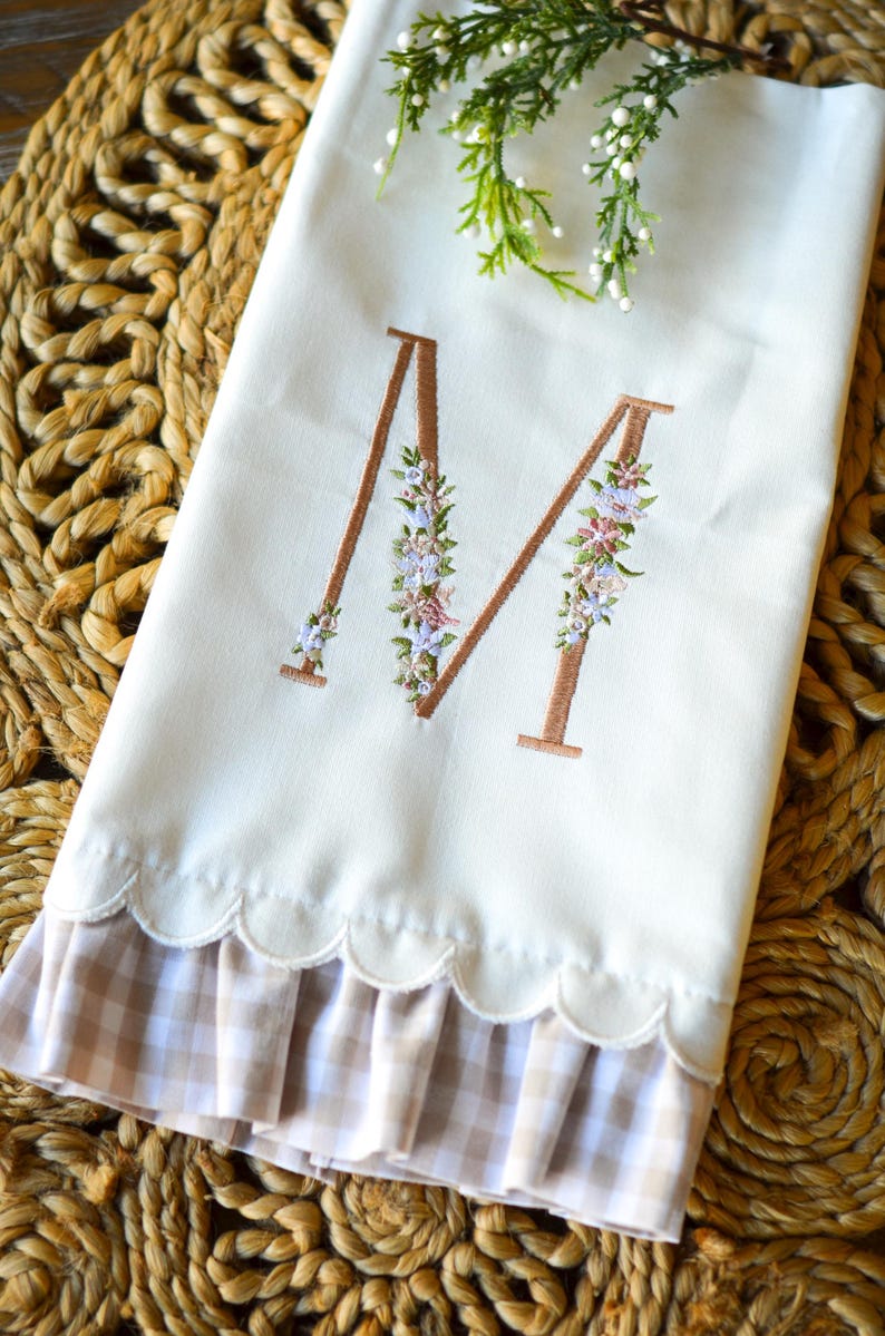 May include: A white hand towel featuring an embroidered letter M in a copper-colored thread, accented with small floral details in shades of pink, blue, and green. The bottom edge has a scalloped finish with a beige and white gingham ruffle trim.