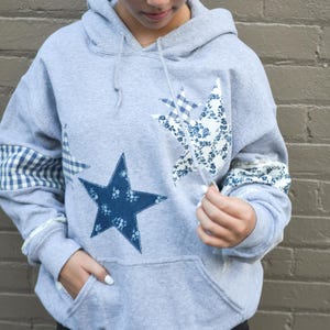 May include: A gray hooded sweatshirt with a large navy blue star and two star-shaped patches. One patch has a blue floral pattern, and the other has a blue and white gingham pattern. The sleeves have matching patterned cuffs.