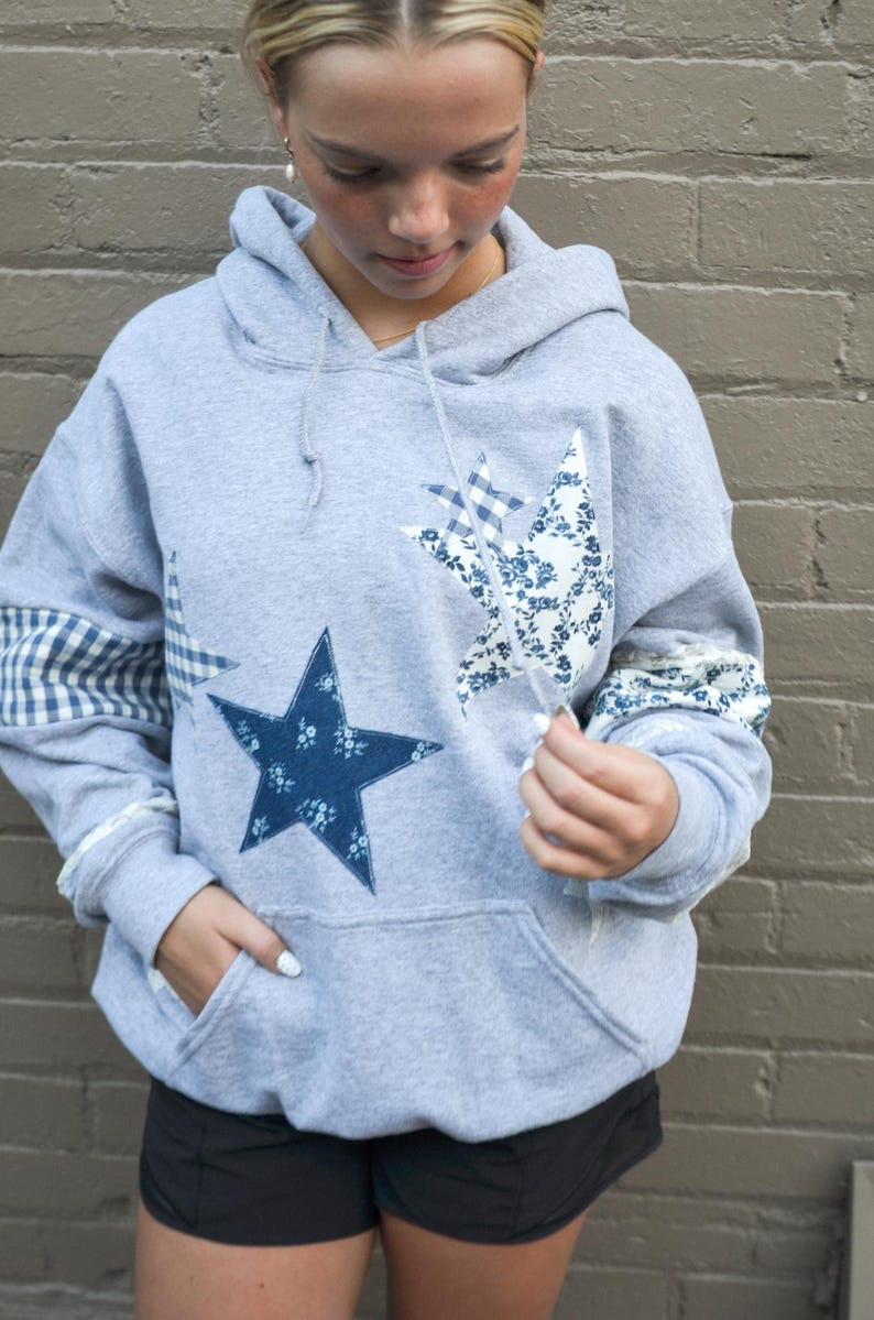 May include: A gray hooded sweatshirt with a large navy blue star and two star-shaped patches. One patch has a blue floral pattern, and the other has a blue and white gingham pattern. The sleeves have matching patterned cuffs.