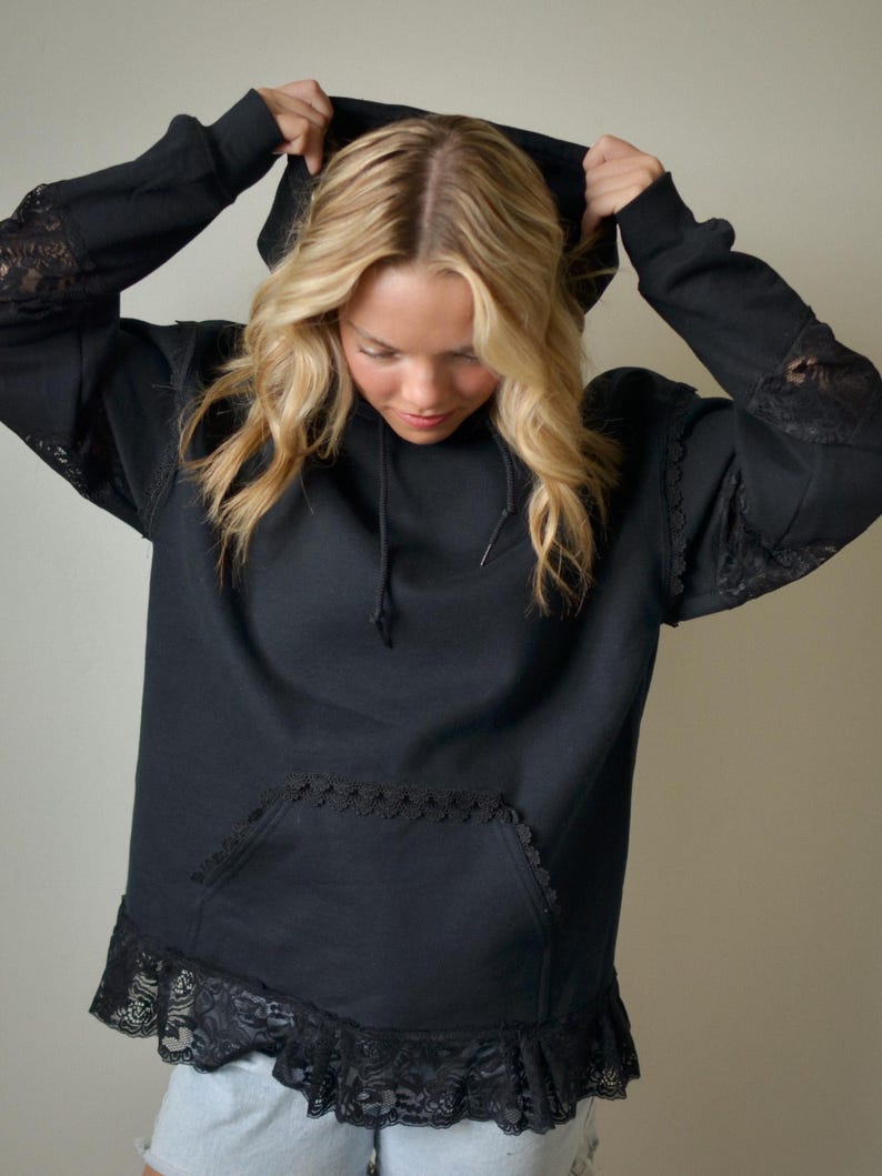 May include: Black hoodie with lace detailing on the sleeves, pocket, and hem. The hoodie has a drawstring hood and a front pocket. The sleeves have lace inserts. The hemline features a wide lace ruffle.