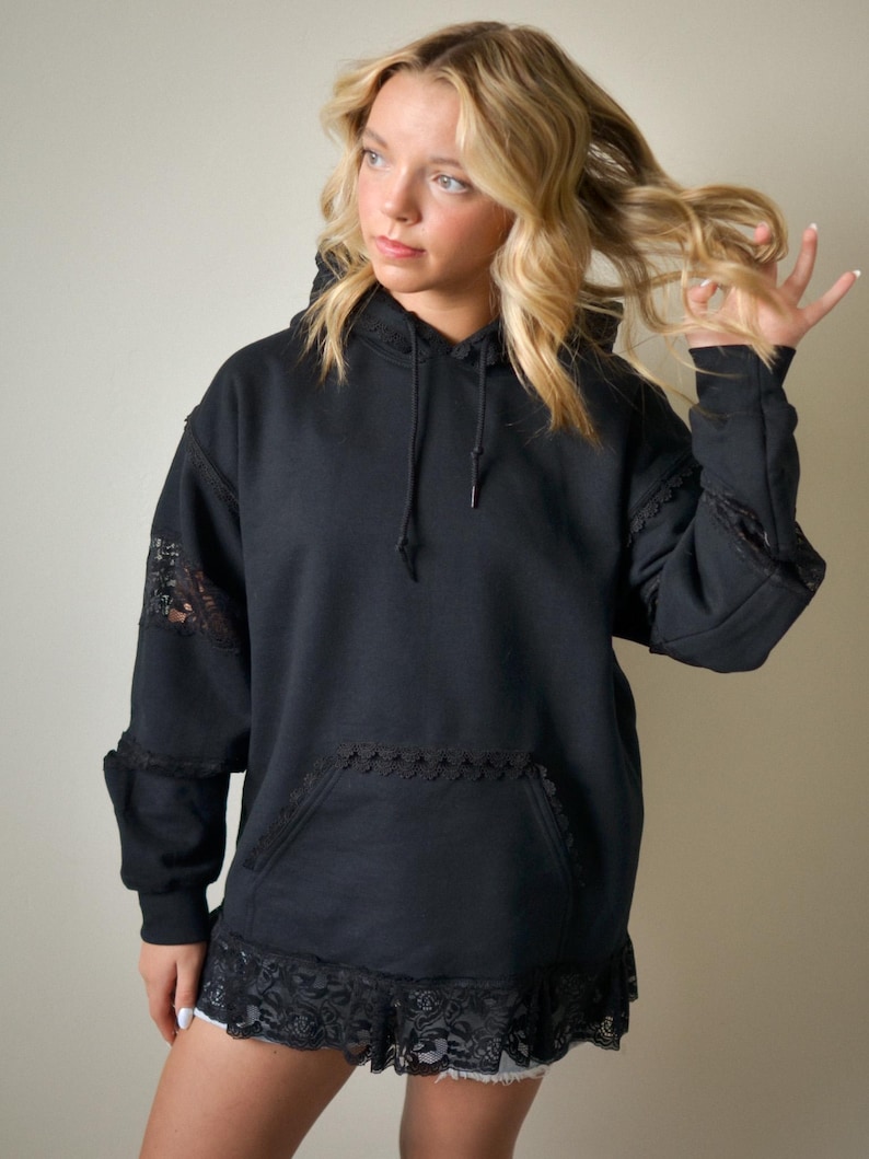 May include: Black hoodie with lace trim on the sleeves, pocket, and hem. The hoodie has a drawstring and a front pocket. The sleeves and hem are adorned with delicate black lace, adding a touch of elegance to the casual design.