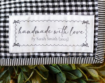 May include: A black and white gingham patterned fabric with a label that reads "handmade with love" by Sarah Smith {2025}. The fabric is trimmed with a black and white striped border. Green leaves and a woven basket are in the foreground.