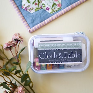 May include: A clear plastic case containing an English paper piecing starter kit. The kit includes various sewing supplies and the text "Cloth & Fable" with the tagline "Every Thread Tells A Story." A floral quilt and pink flowers are also visible.