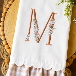 May include: A white hand towel featuring a large embroidered letter M in a copper-colored thread, decorated with small pink, purple, and green floral accents. The bottom edge has a scalloped hem and a ruffled light brown and white gingham trim.