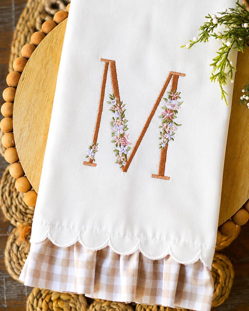 May include: A white hand towel featuring a large embroidered letter M in a copper-colored thread, decorated with small pink, purple, and green floral accents. The bottom edge has a scalloped hem and a ruffled light brown and white gingham trim.