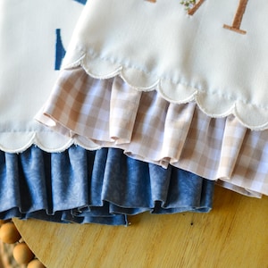 May include: Two off-white fabric towels with scalloped edges and ruffled trim. One towel features a brown embroidered letter M decorated with small floral accents, while the other displays a partial blue letter S. The ruffles are beige gingham.